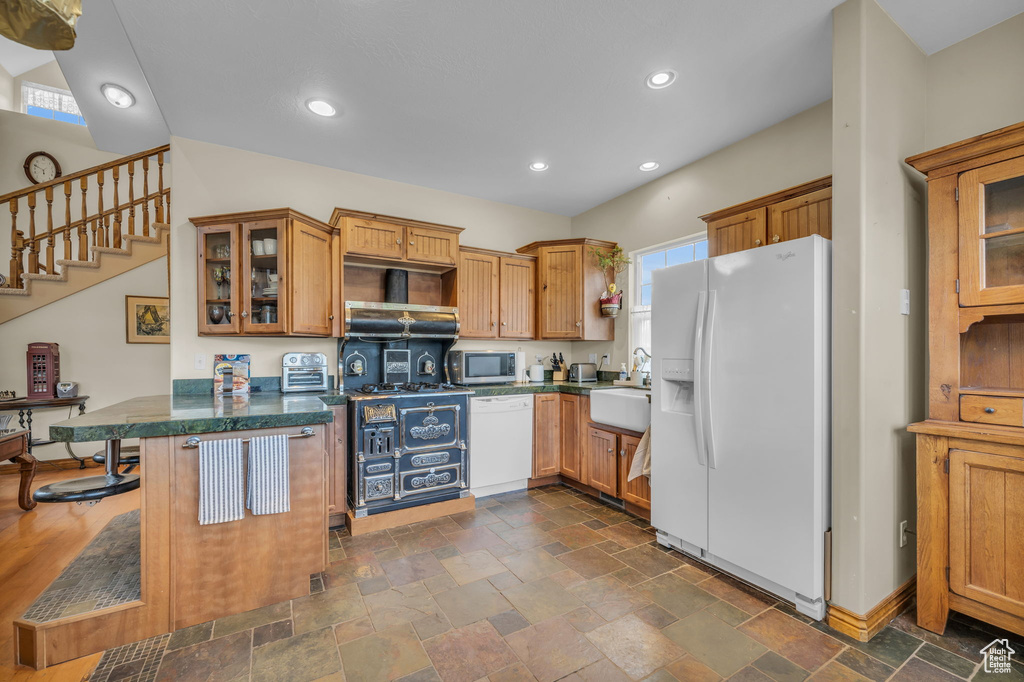 Kitchen with white appliances, kitchen peninsula, wall chimney exhaust hood, sink, and dark tile flooring