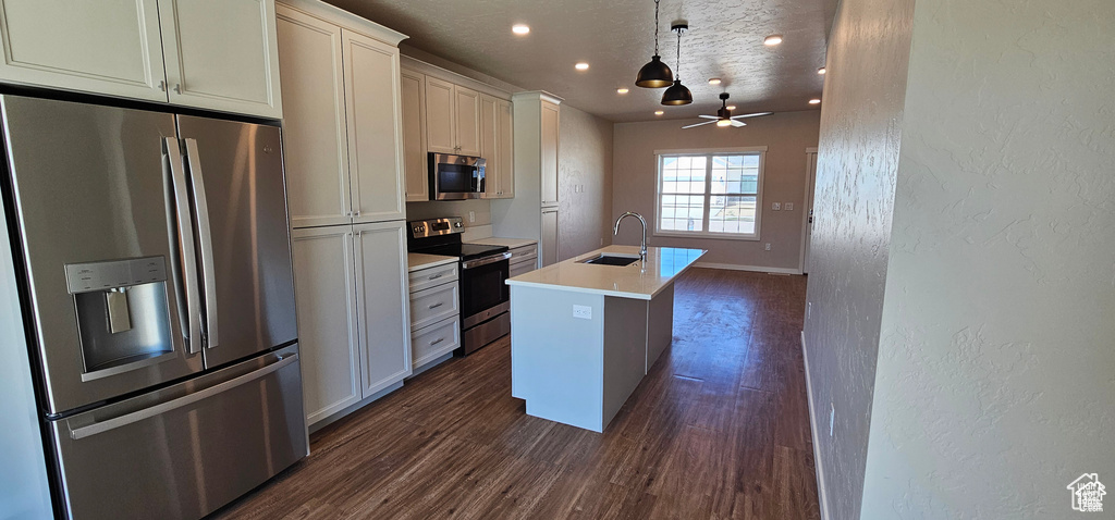 Kitchen featuring ceiling fan, stainless steel appliances, pendant lighting, dark wood-type flooring, and a kitchen island with sink