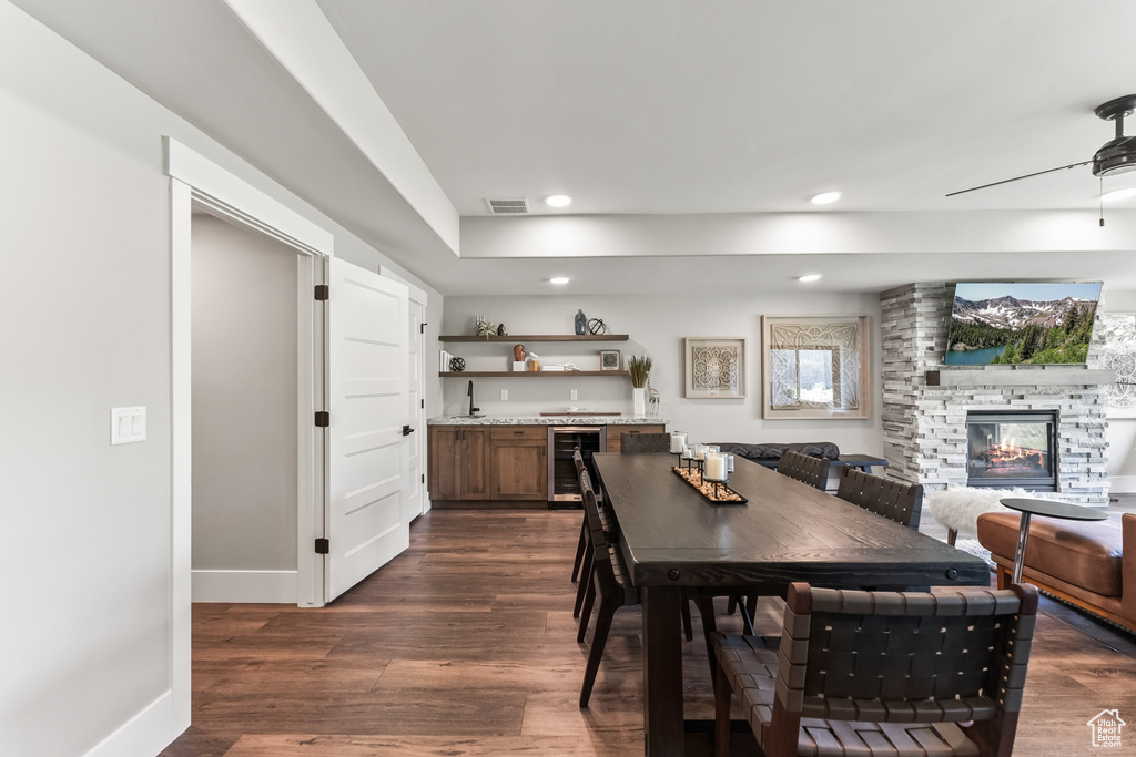 Dining space featuring wine cooler, ceiling fan, a stone fireplace, and dark wood-type flooring