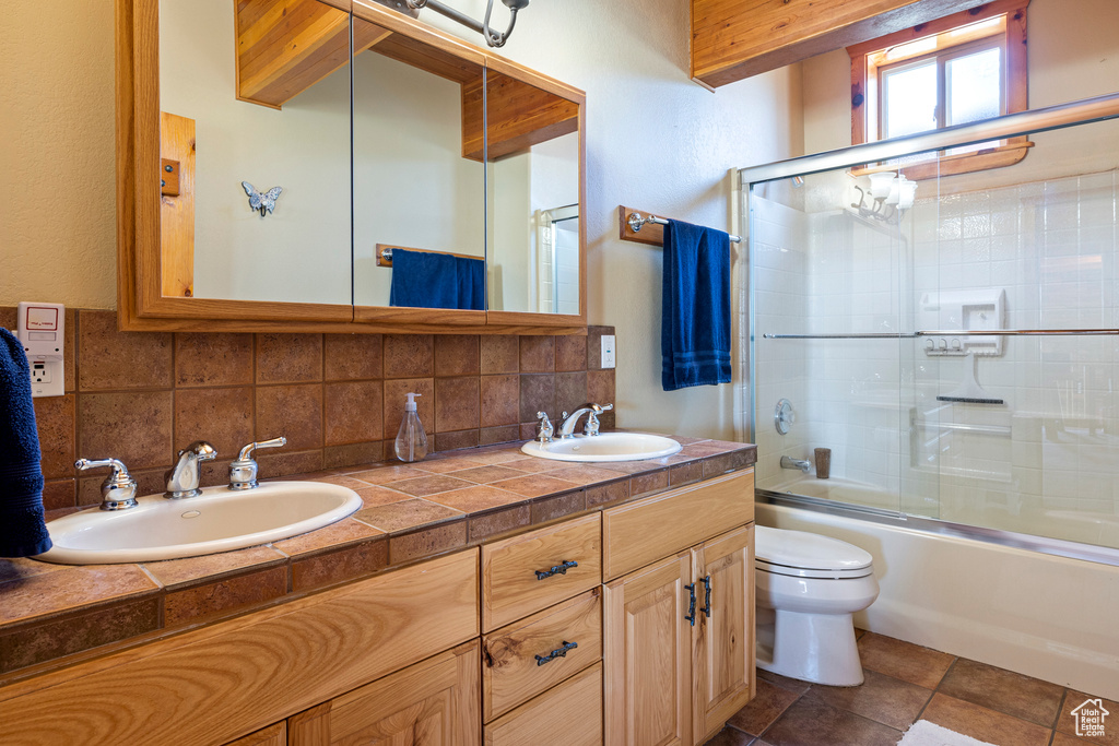 Full bathroom with backsplash, dual vanity, enclosed tub / shower combo, toilet, and tile flooring