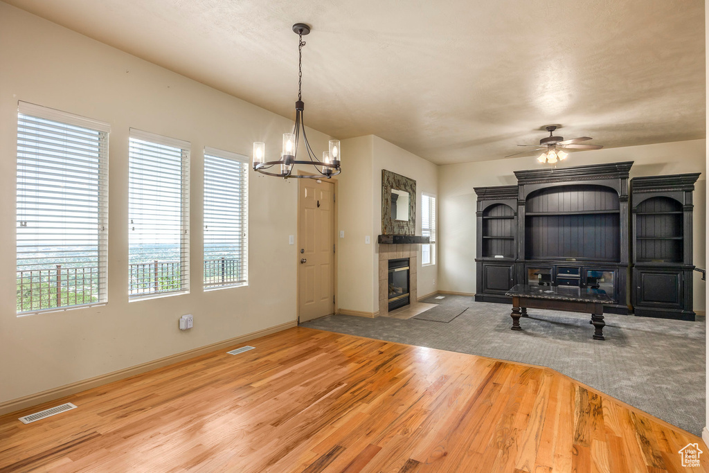 Unfurnished living room featuring wood-type flooring, a tiled fireplace, and ceiling fan with notable chandelier