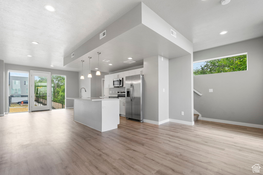 Kitchen featuring light hardwood / wood-style floors, stainless steel appliances, hanging light fixtures, white cabinets, and a kitchen island with sink