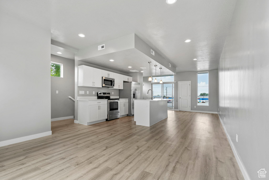 Kitchen with an island with sink, stainless steel appliances, decorative light fixtures, light wood-type flooring, and white cabinets