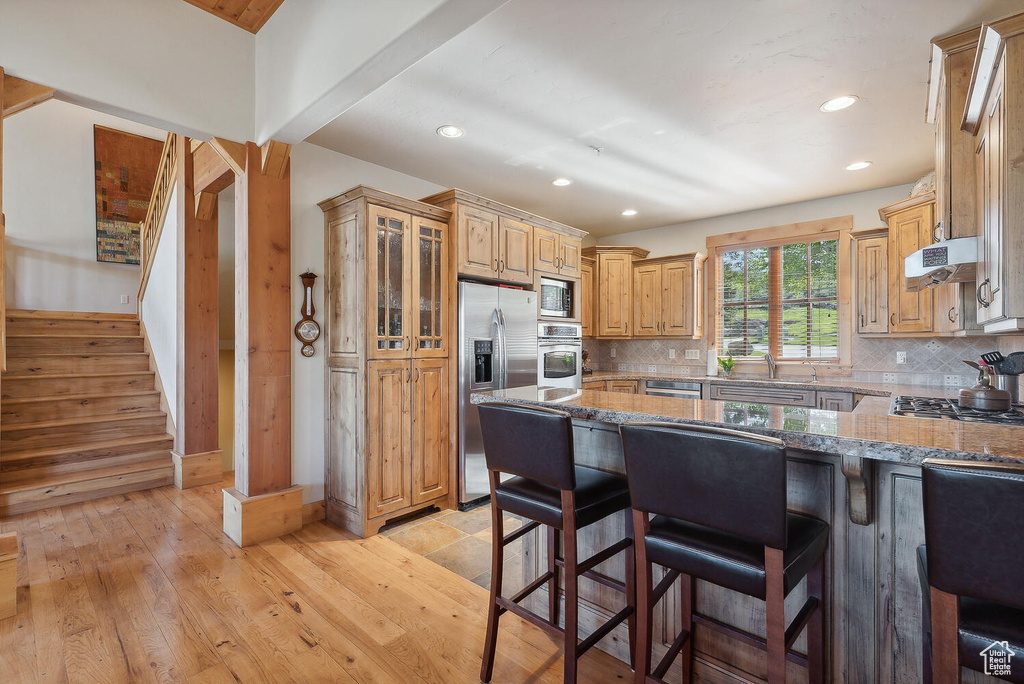 Kitchen featuring tasteful backsplash, wall chimney exhaust hood, light wood-type flooring, appliances with stainless steel finishes, and a breakfast bar