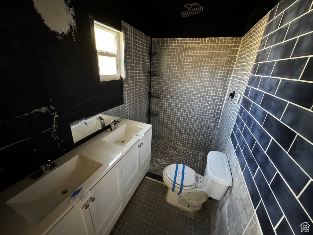 Bathroom featuring double sink, tile floors, tile walls, vanity with extensive cabinet space, and toilet