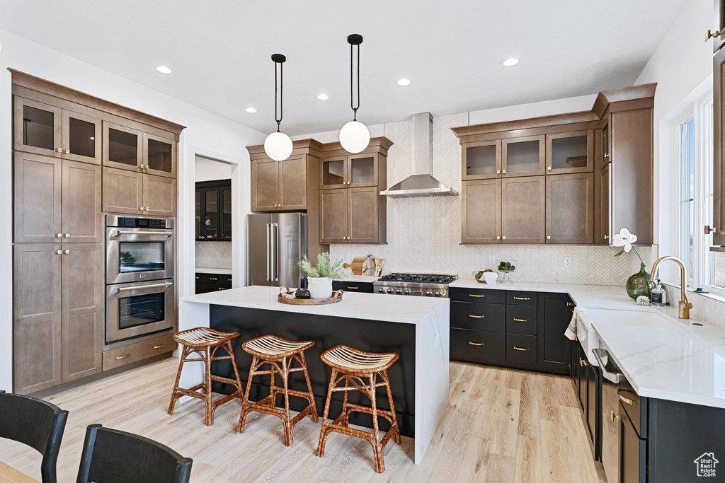 Kitchen featuring a kitchen island, light hardwood / wood-style flooring, stainless steel appliances, wall chimney exhaust hood, and backsplash