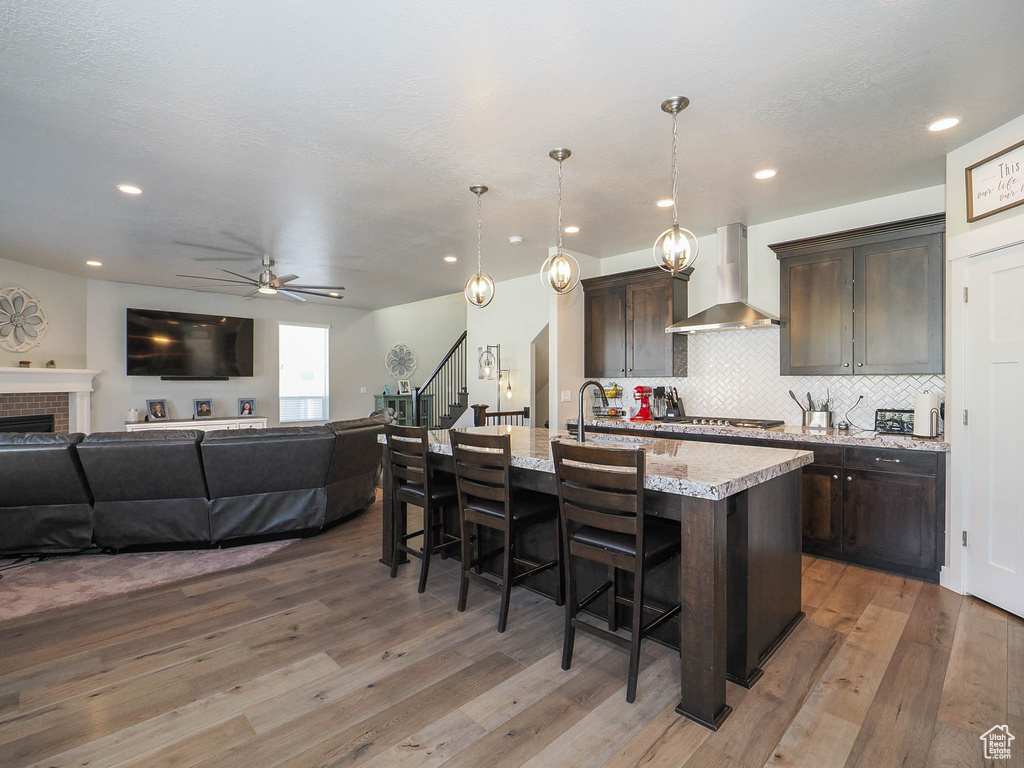 Kitchen with wall chimney range hood, a fireplace, a kitchen island with sink, and wood-type flooring