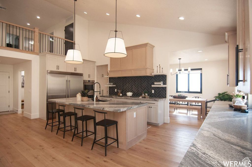 Kitchen featuring decorative light fixtures, tasteful backsplash, a kitchen island with sink, light wood-type flooring, and high vaulted ceiling