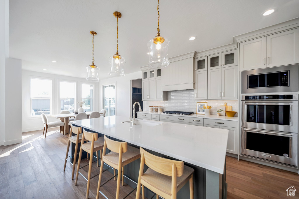 Kitchen featuring appliances with stainless steel finishes, hardwood / wood-style floors, hanging light fixtures, decorative backsplash, and an island with sink