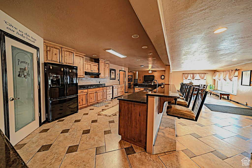 Kitchen with a breakfast bar area, light tile flooring, black fridge, a textured ceiling, and a kitchen island with sink