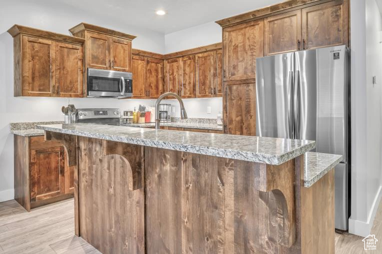 Kitchen featuring light wood-type flooring, stainless steel appliances, sink, light stone counters, and a center island with sink