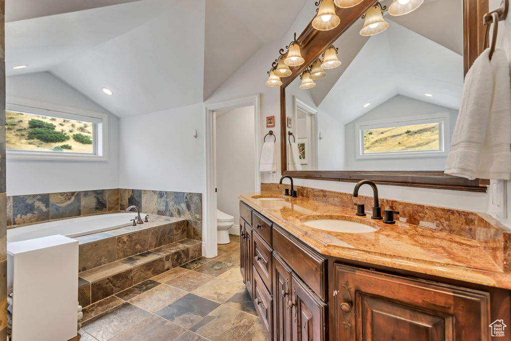 Bathroom featuring toilet, a relaxing tiled tub, vanity, and vaulted ceiling