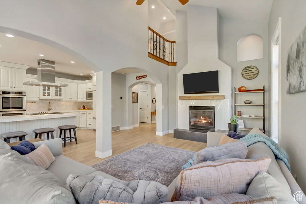 Living room with a towering ceiling, a tiled fireplace, light wood-type flooring, and ceiling fan