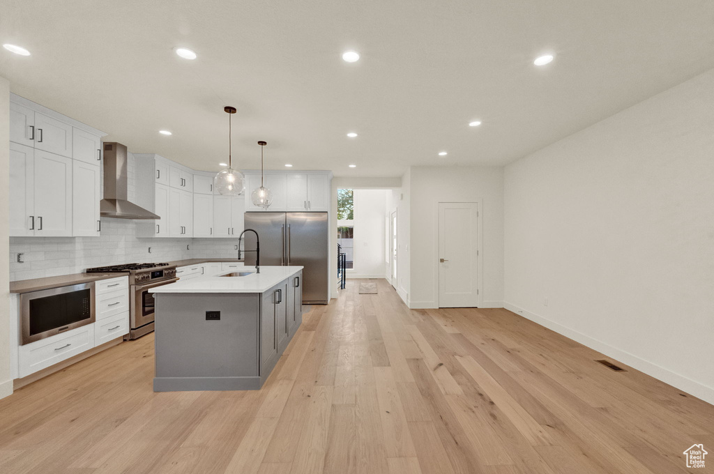 Kitchen featuring wall chimney range hood, an island with sink, built in appliances, and light wood-type flooring