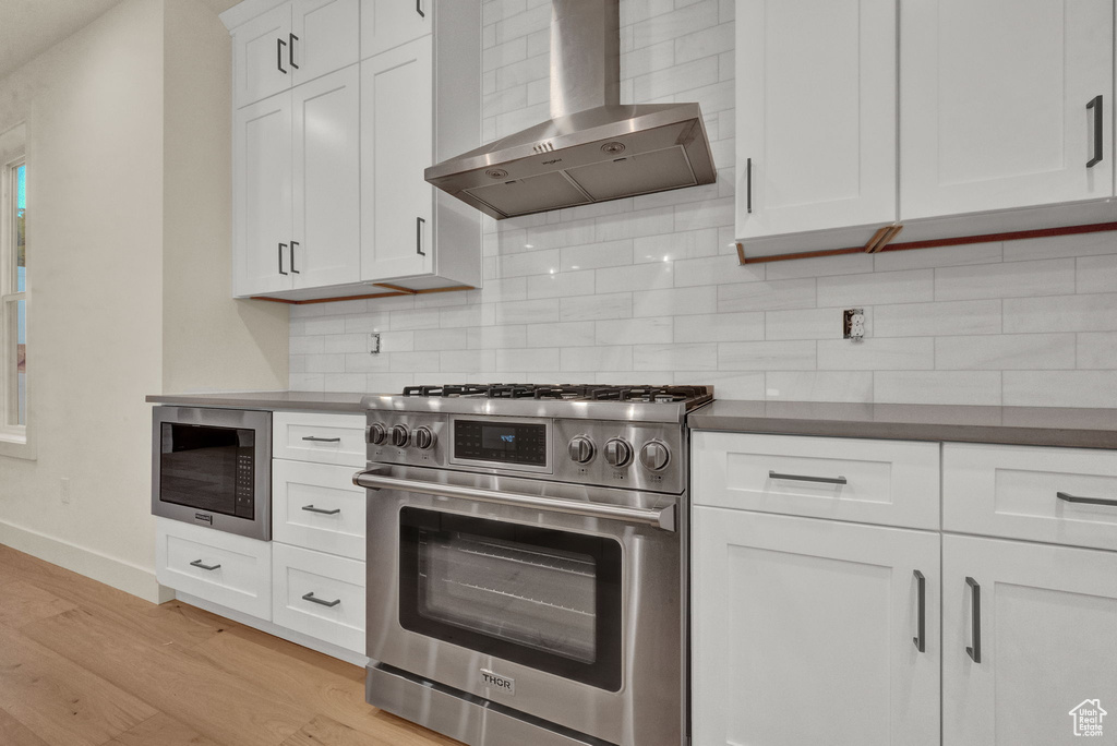 Kitchen with white cabinetry, light wood-type flooring, stainless steel appliances, wall chimney exhaust hood, and tasteful backsplash