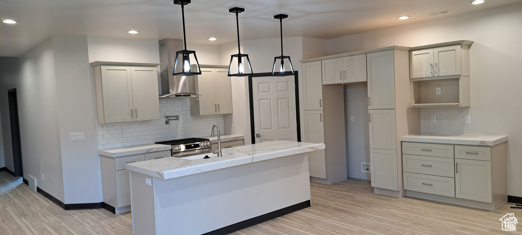 Kitchen featuring light hardwood / wood-style flooring, a center island with sink, stainless steel range with gas cooktop, pendant lighting, and wall chimney exhaust hood