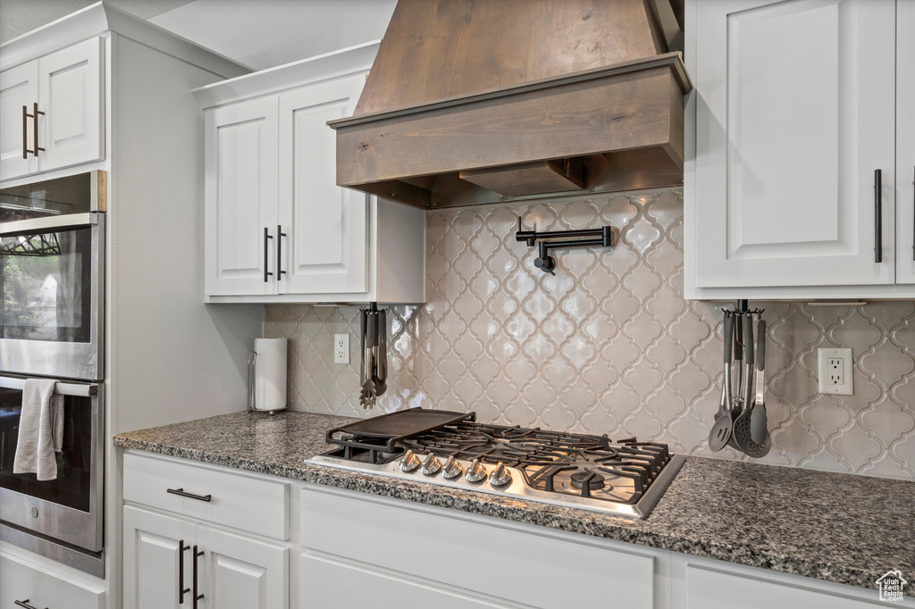 Kitchen with white cabinetry, dark stone countertops, custom range hood, stainless steel appliances, and backsplash