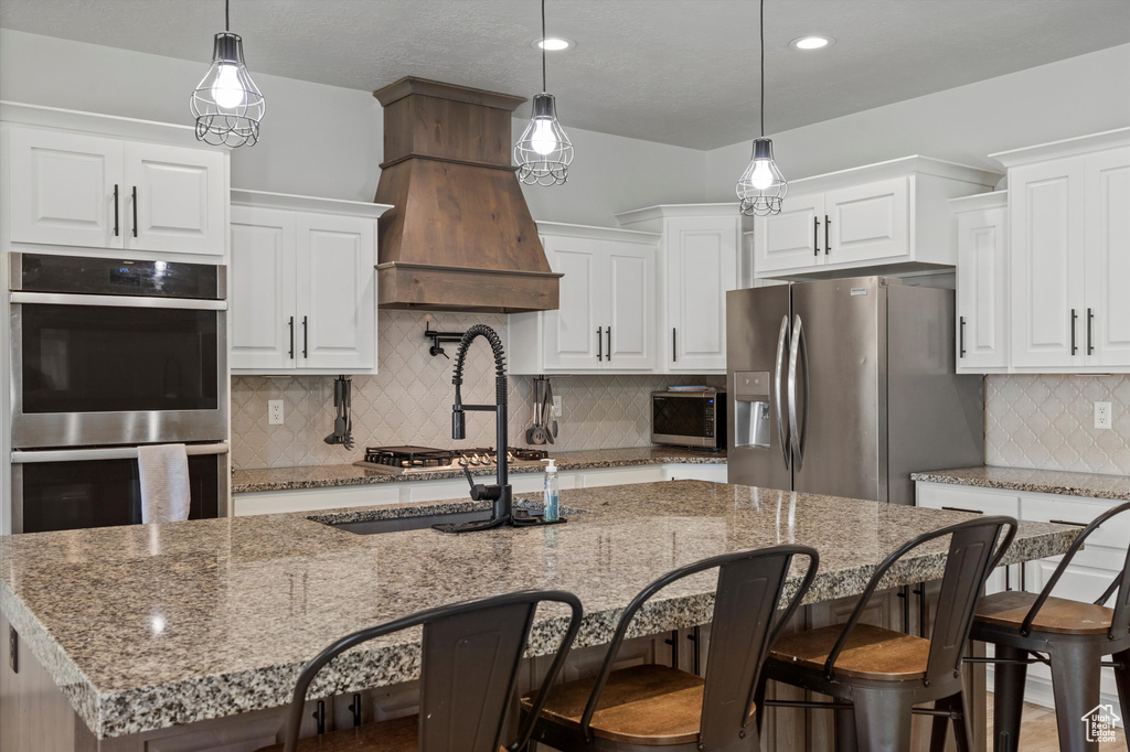 Kitchen featuring tasteful backsplash, a center island with sink, custom range hood, white cabinetry, and appliances with stainless steel finishes