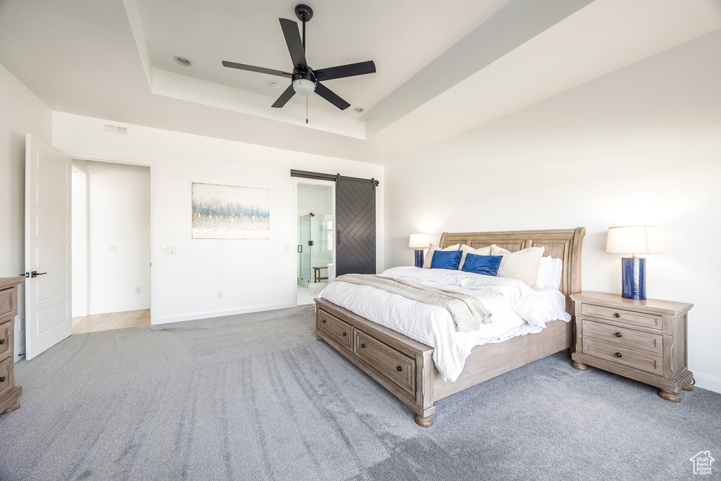 Carpeted bedroom featuring a barn door, ceiling fan, a tray ceiling, and ensuite bathroom