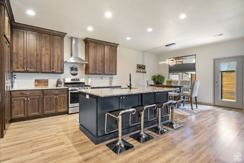 Kitchen with light hardwood / wood-style floors, stainless steel range, an island with sink, wall chimney exhaust hood, and sink