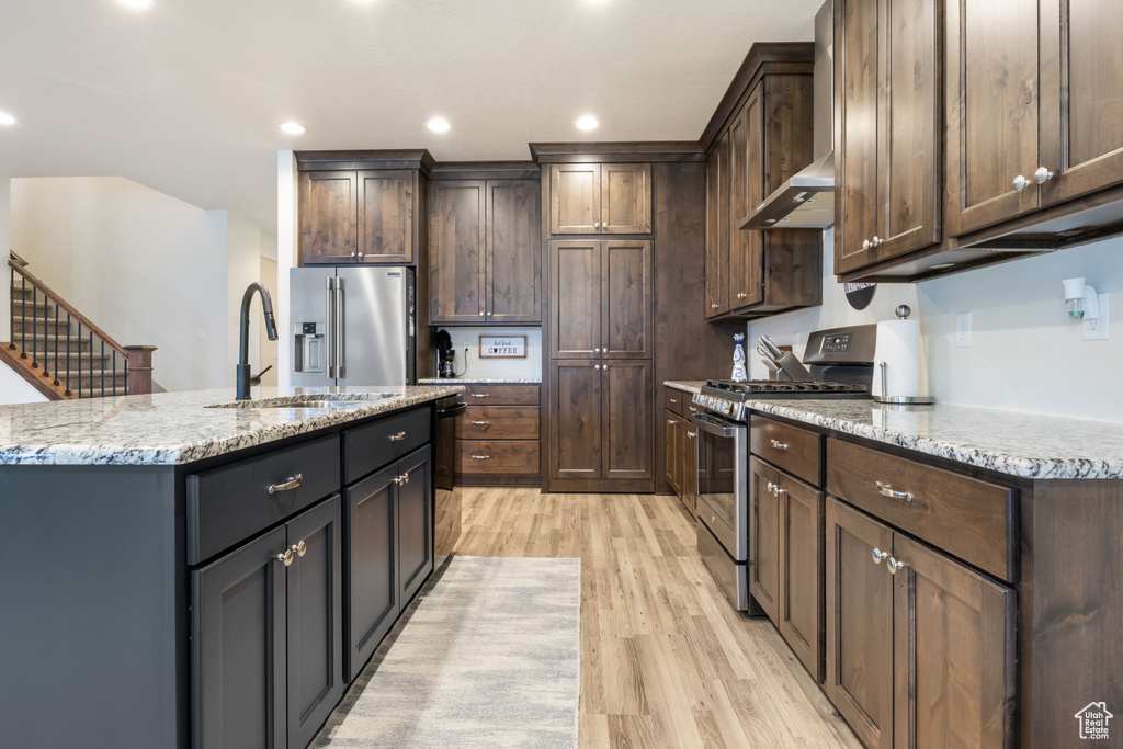 Kitchen with light stone countertops, light hardwood / wood-style flooring, stainless steel appliances, a kitchen island with sink, and dark brown cabinets