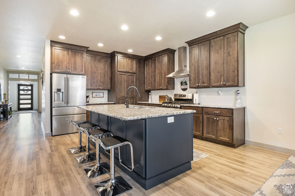 Kitchen with light hardwood / wood-style flooring, a center island with sink, wall chimney range hood, sink, and appliances with stainless steel finishes