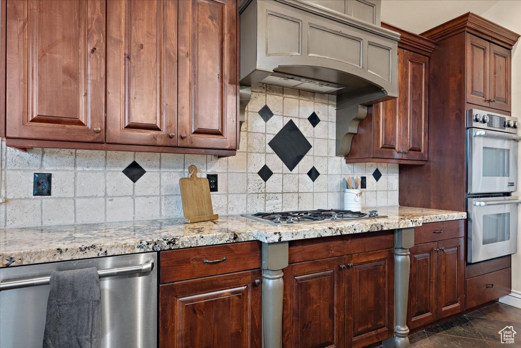 Kitchen with light stone counters, custom range hood, backsplash, dark tile flooring, and appliances with stainless steel finishes