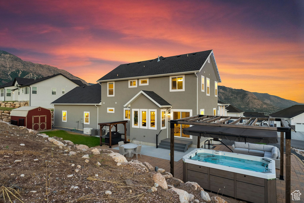 Back house at dusk featuring a pergola, a storage unit, a patio, a mountain view, and a hot tub