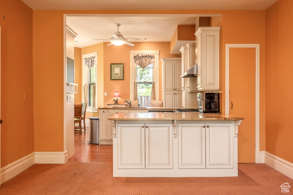 Kitchen featuring wall chimney range hood, ceiling fan, kitchen peninsula, and light stone counters