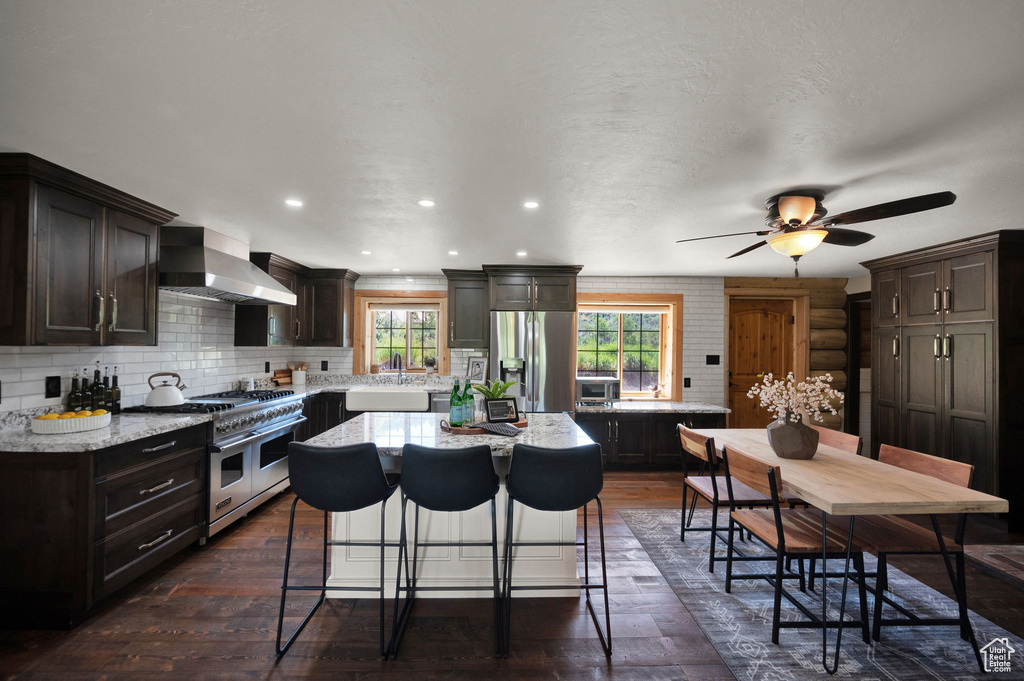 Kitchen with dark hardwood / wood-style floors, ceiling fan, stainless steel appliances, a kitchen island, and wall chimney range hood