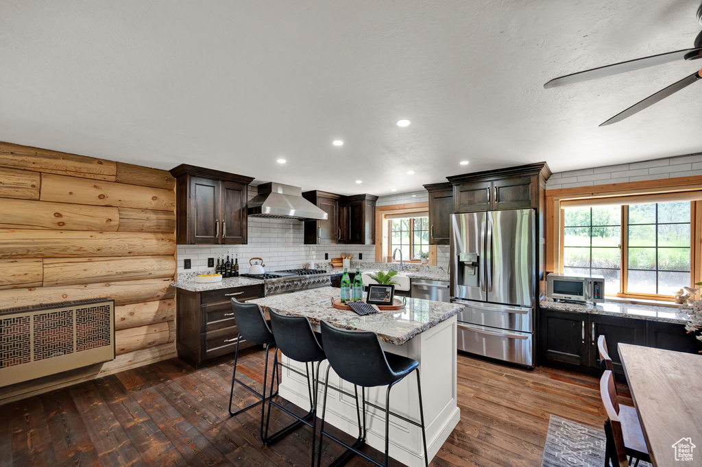 Kitchen featuring dark wood-type flooring, a kitchen island, stainless steel appliances, wall chimney exhaust hood, and ceiling fan