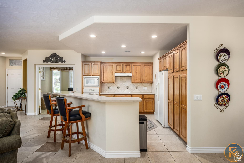 Kitchen with light tile flooring, backsplash, a breakfast bar area, a center island, and white appliances