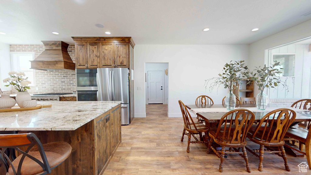 Kitchen with light stone countertops, light hardwood / wood-style flooring, custom exhaust hood, stainless steel appliances, and a breakfast bar