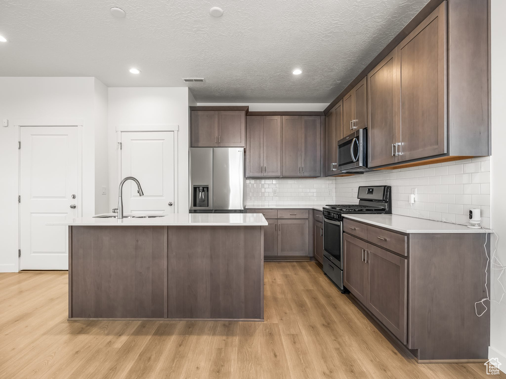 Kitchen with backsplash, appliances with stainless steel finishes, light hardwood / wood-style floors, and an island with sink