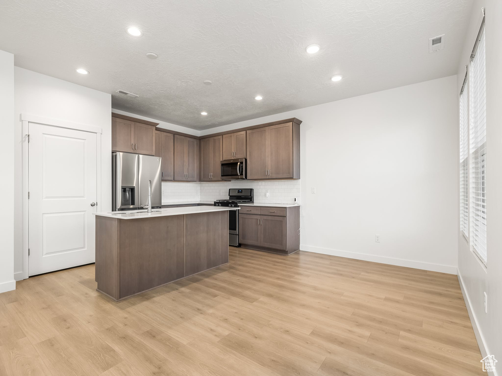 Kitchen with appliances with stainless steel finishes, light wood-type flooring, tasteful backsplash, and an island with sink