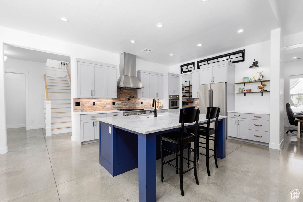 Kitchen with wall chimney range hood, appliances with stainless steel finishes, white cabinets, and a kitchen island with sink