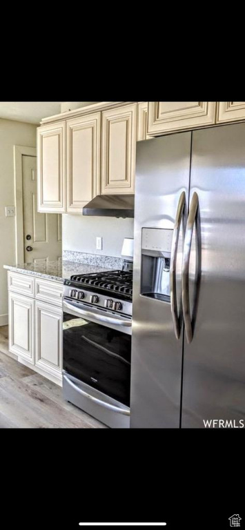 Kitchen featuring stainless steel appliances, light hardwood / wood-style floors, light stone counters, cream cabinets, and wall chimney exhaust hood