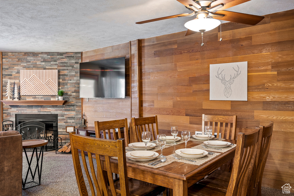 Carpeted dining room featuring ceiling fan, a stone fireplace, a textured ceiling, and wooden walls