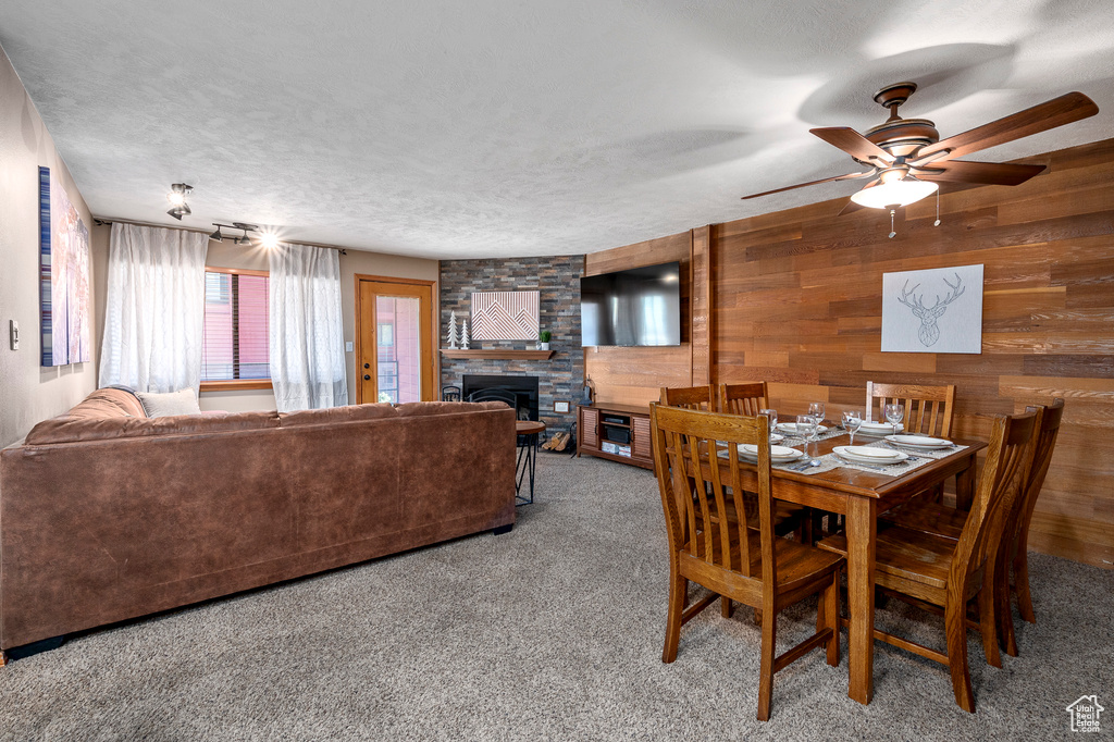 Carpeted dining area featuring a textured ceiling, a stone fireplace, wooden walls, and ceiling fan