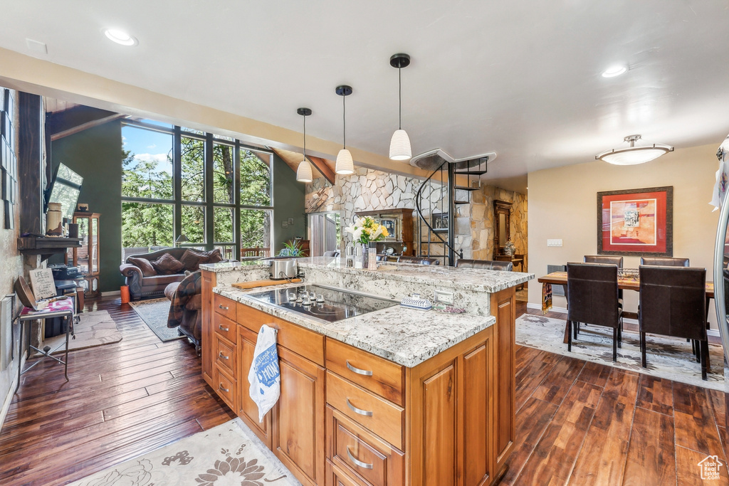 Kitchen with light stone countertops, a center island with sink, black electric cooktop, pendant lighting, and dark wood-type flooring