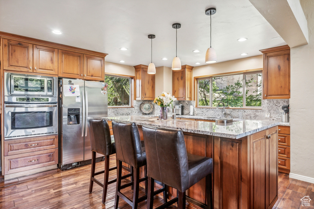 Kitchen with light stone countertops, stainless steel appliances, hardwood / wood-style flooring, decorative light fixtures, and tasteful backsplash