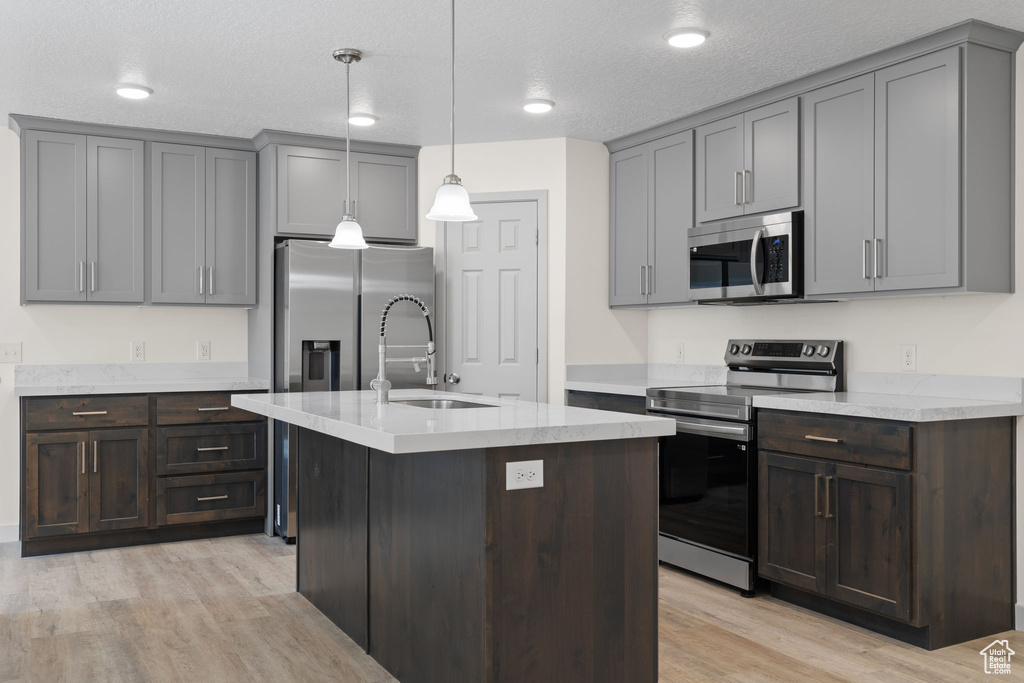 Kitchen featuring appliances with stainless steel finishes, an island with sink, a textured ceiling, hanging light fixtures, and light hardwood / wood-style floors