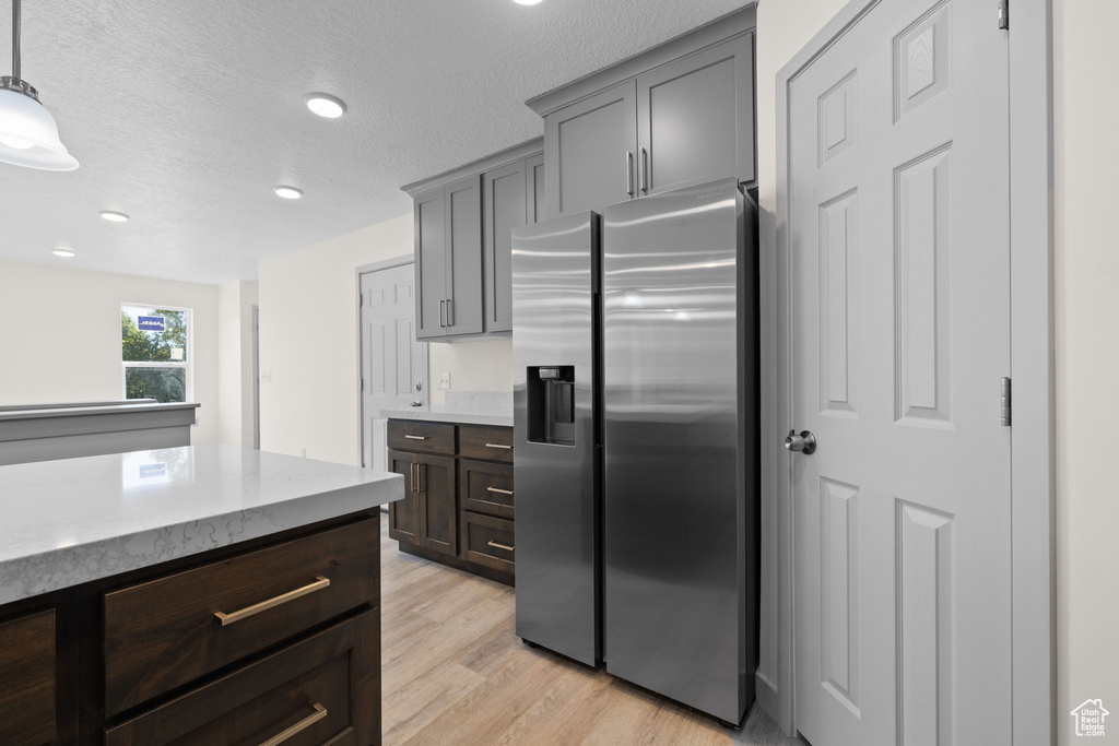 Kitchen featuring light wood-type flooring, a textured ceiling, stainless steel fridge, decorative light fixtures, and light stone counters