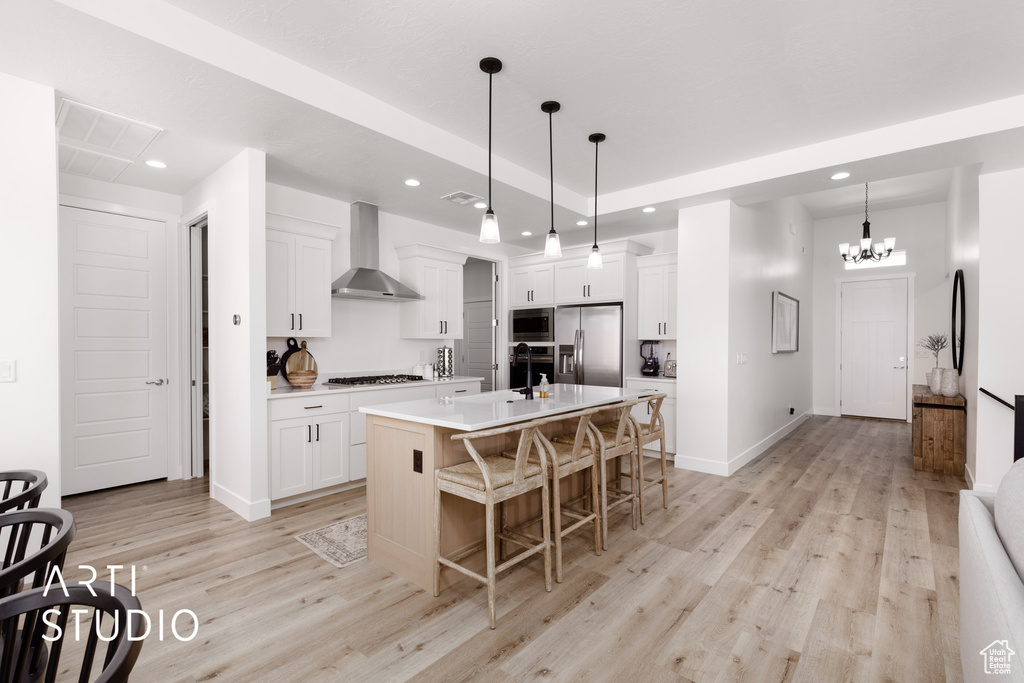 Kitchen with wall chimney range hood, pendant lighting, light wood-type flooring, appliances with stainless steel finishes, and a kitchen island with sink