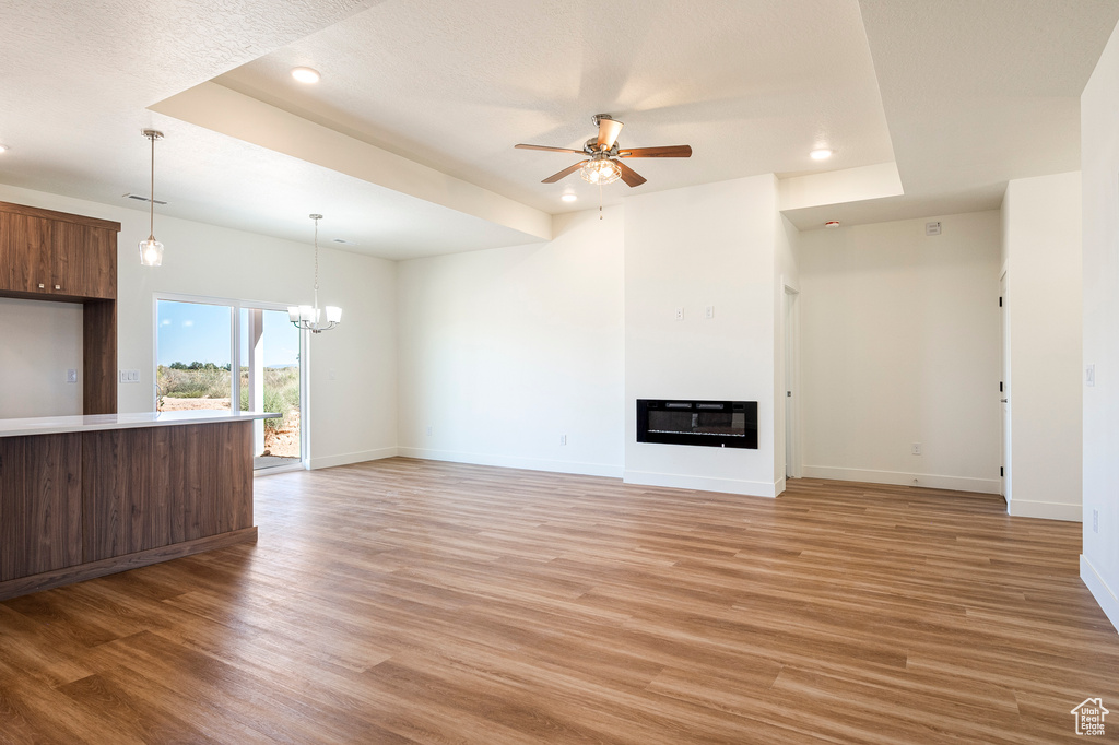 Unfurnished living room with ceiling fan with notable chandelier, a textured ceiling, a tray ceiling, and light hardwood / wood-style floors