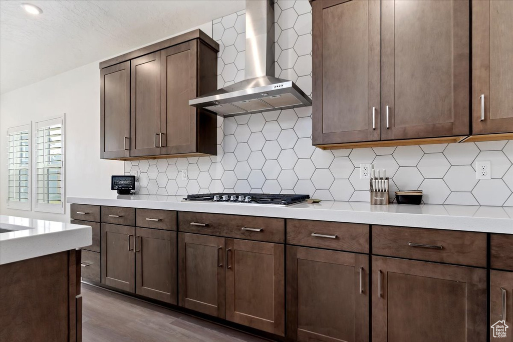 Kitchen featuring stainless steel gas stovetop, light wood-type flooring, wall chimney range hood, backsplash, and dark brown cabinetry