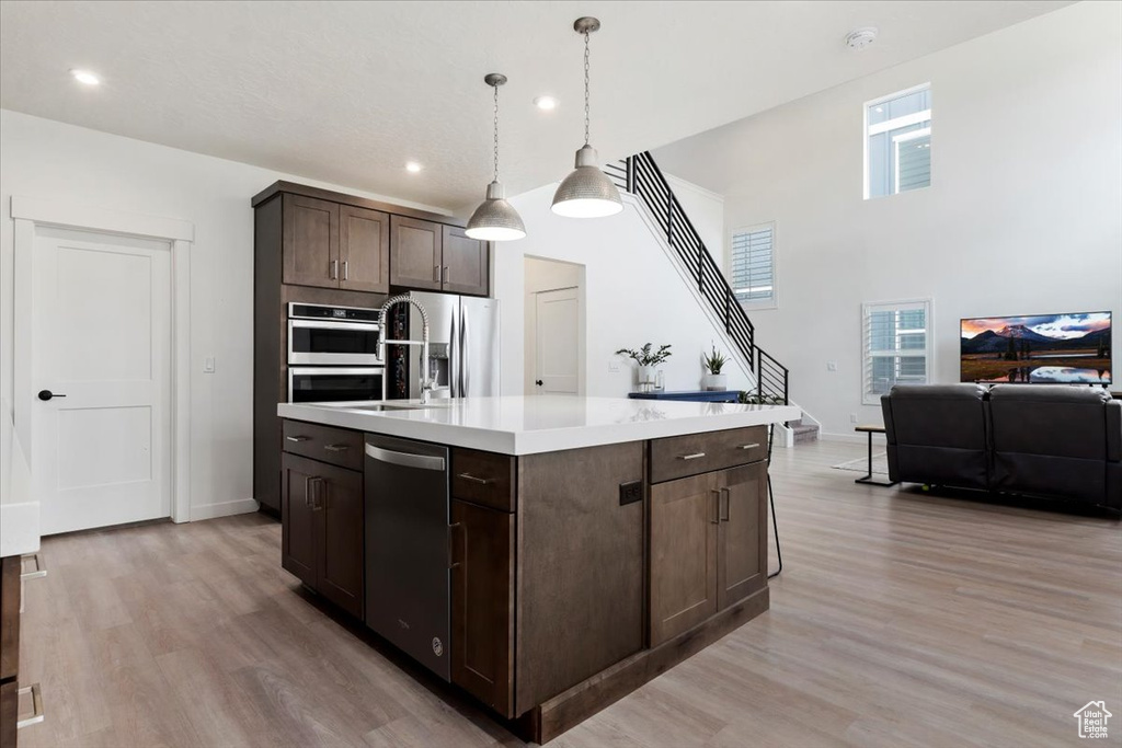 Kitchen with appliances with stainless steel finishes, a center island with sink, dark brown cabinets, decorative light fixtures, and light wood-type flooring