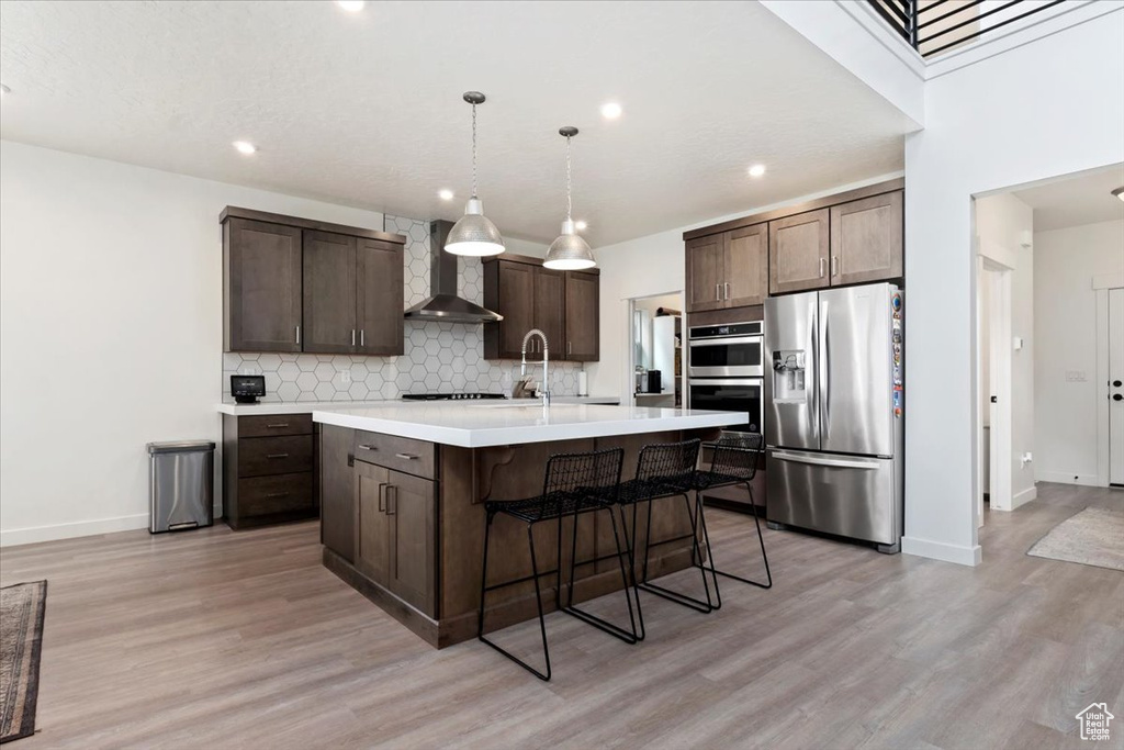 Kitchen with appliances with stainless steel finishes, light hardwood / wood-style flooring, decorative backsplash, an island with sink, and wall chimney exhaust hood