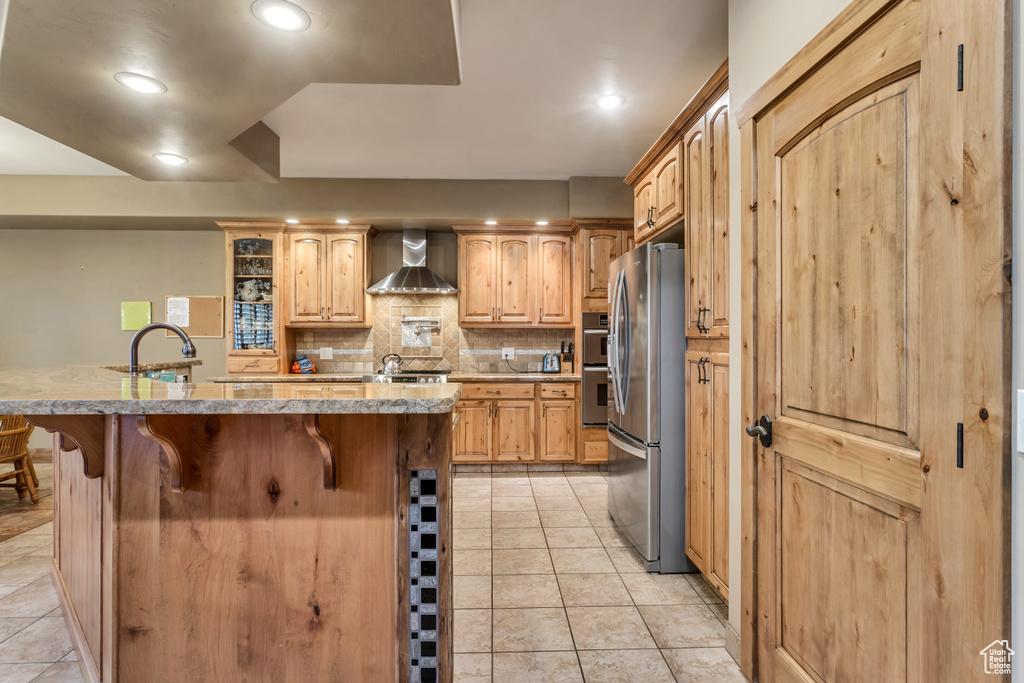 Kitchen with light tile patterned floors, wall chimney range hood, decorative backsplash, stainless steel fridge, and a kitchen bar