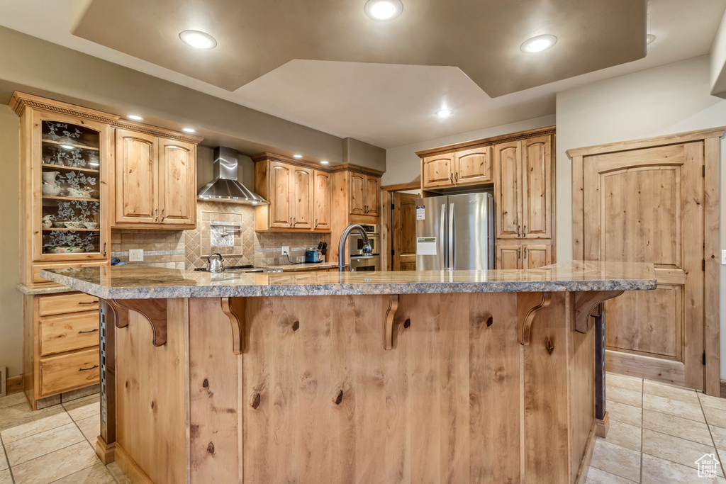 Kitchen with light tile patterned flooring, wall chimney exhaust hood, appliances with stainless steel finishes, a kitchen breakfast bar, and backsplash